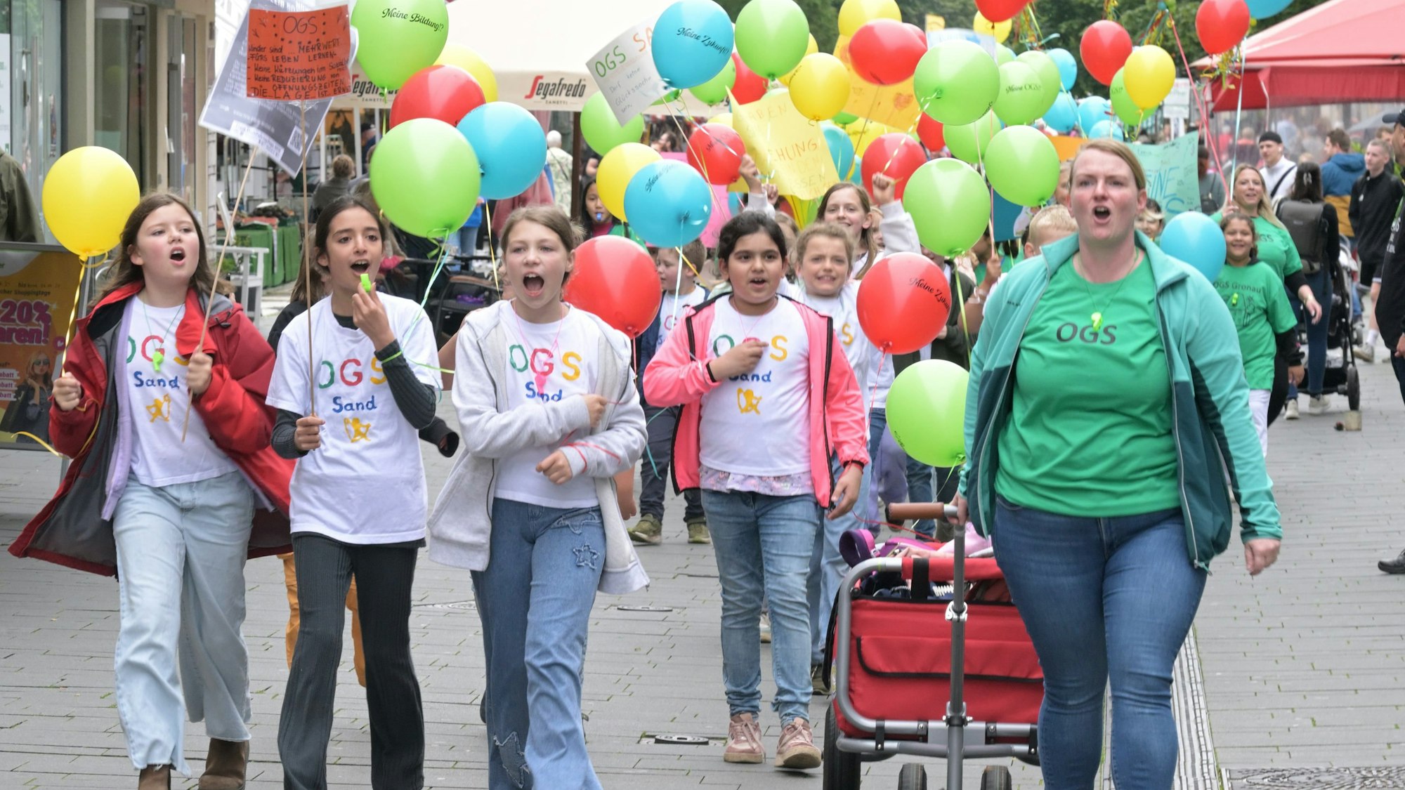 Im Vordergrund sind vier Mädchen mit Trillerpfeifen und eine Erzieherin mit Bollerwagen zu sehen. Im Hintergrund sind vor allem die Luftballons zu sehen, die die Kinder in der Hand halten.