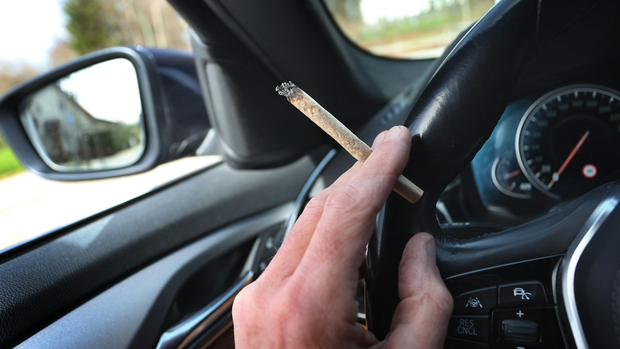 Ein Mann sitzt mit einem Joint zwischen den Fingern am Steuer eines Autos.