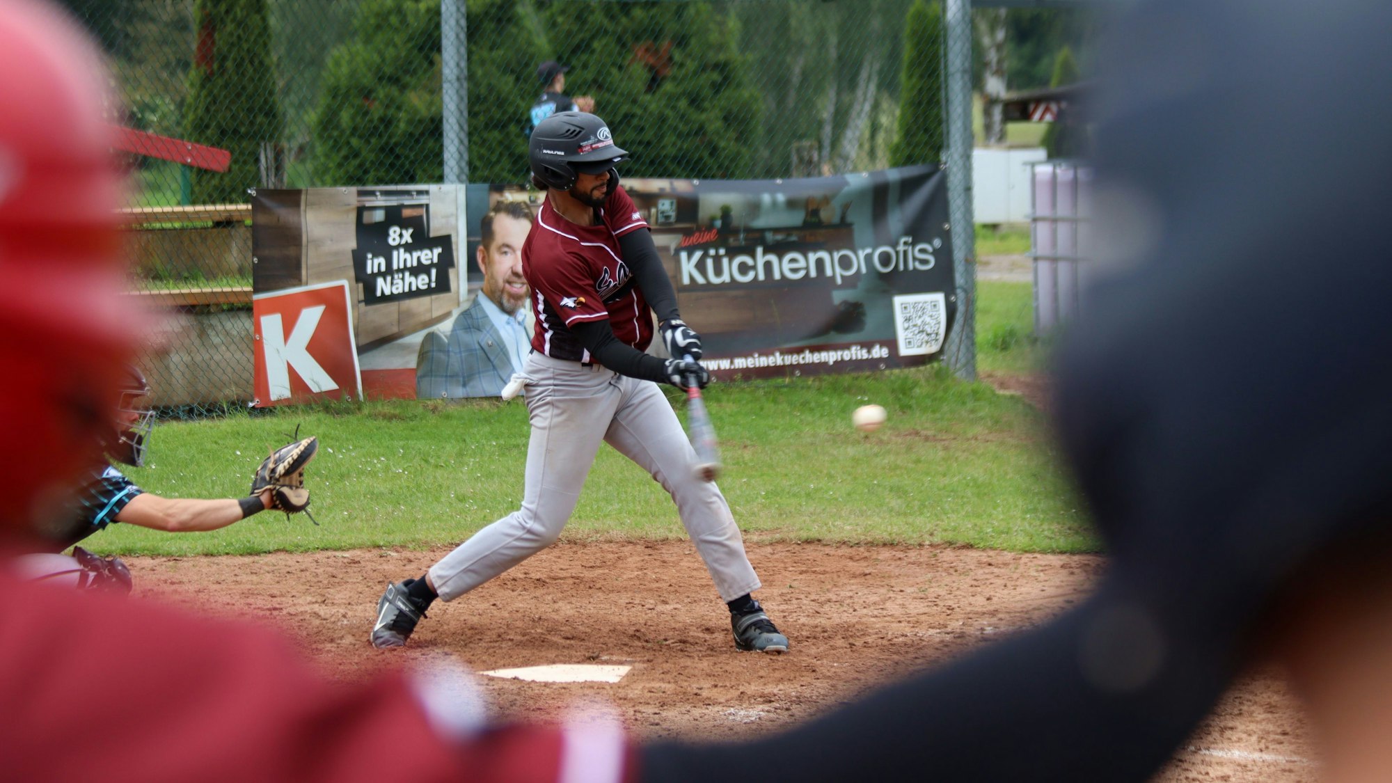 Ein Baseballer trifft einen Ball. Im Vordergrund unscharf seine Mitspieler, die zuschauen.