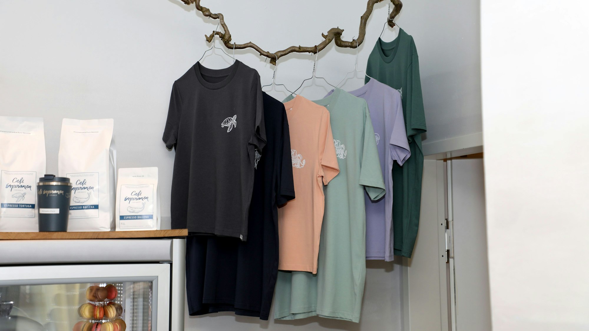 T-Shirts mit wal-Emblem hängen an einem gewundenen Stock im Café Esperanza in Rösrath-Forsbach. Daneben sind der hauseigene Kaffee und ein Kaffeebecher zu sehen.