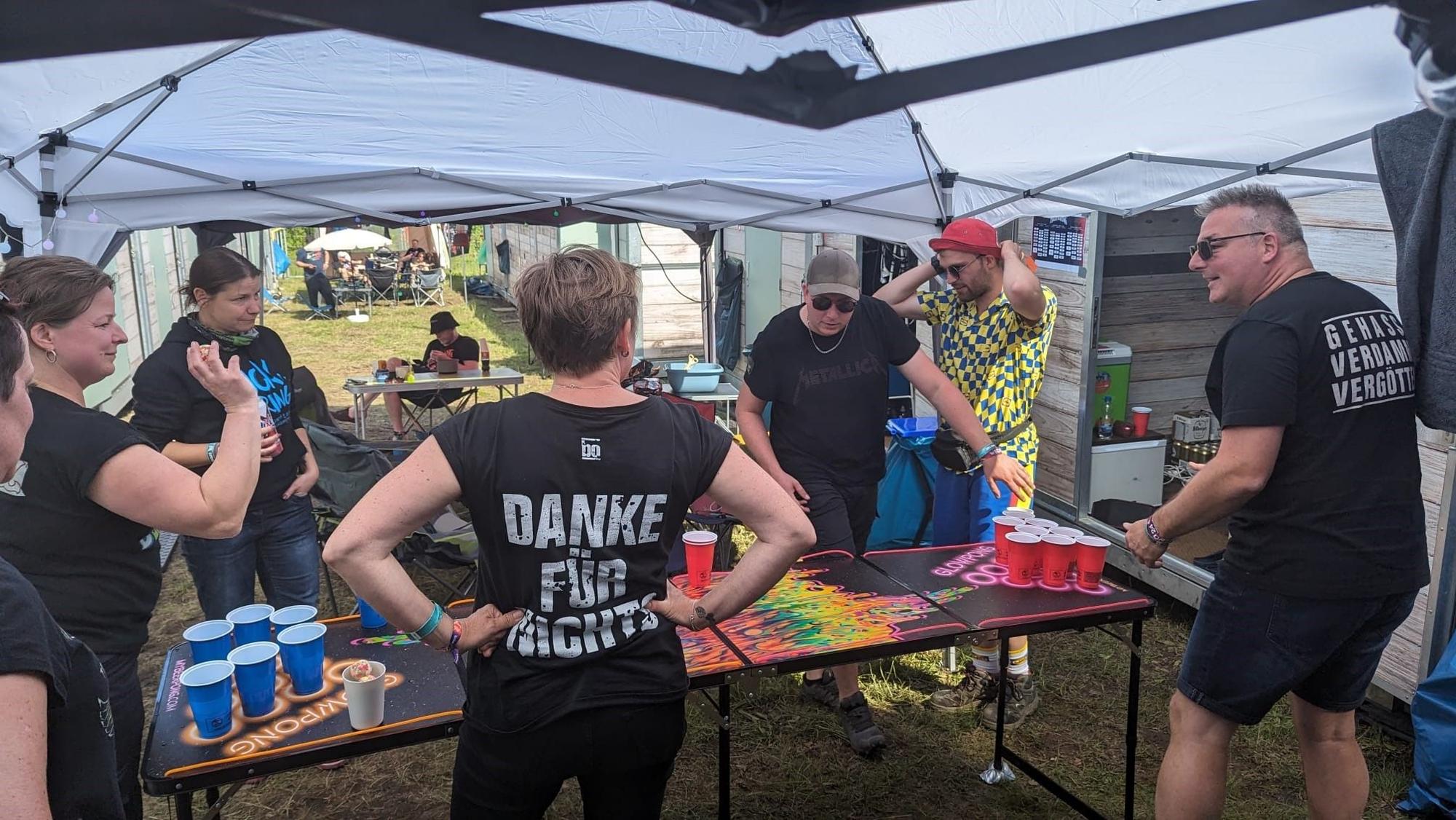 Eine Gruppe von Menschen steht an einem Beer-Pong-Tisch unter einem Pavillon auf einem Camping-Platz.