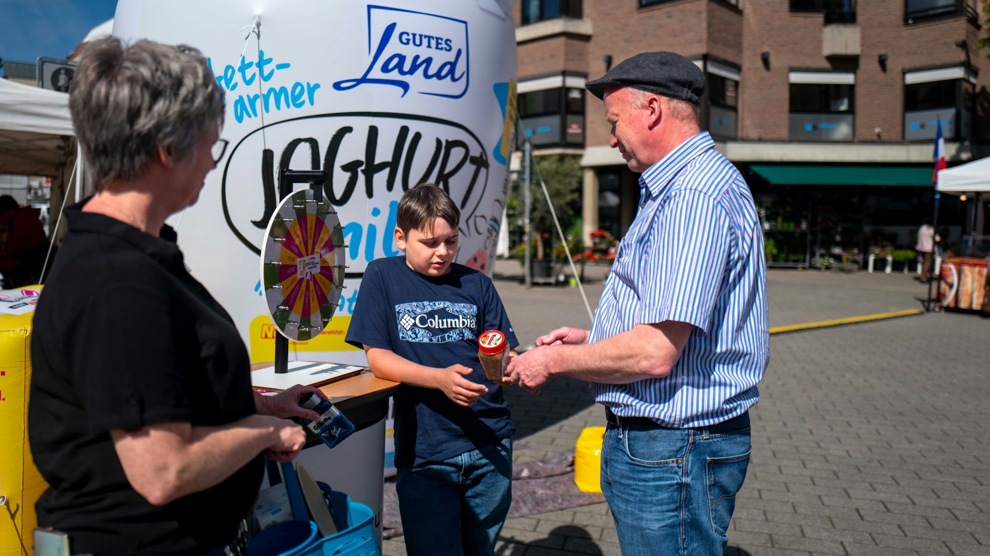 Ein Junge steht an einem kleinen Glücksrad, dass vor einem übergroßen Joghurtbecher aufgestellt wurde und unterhält sich mit einem Mann und einer Frau.