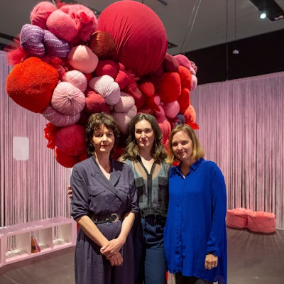 Drei Frauen stehen vor einem großen Gebilde aus roten Stoffkugeln.