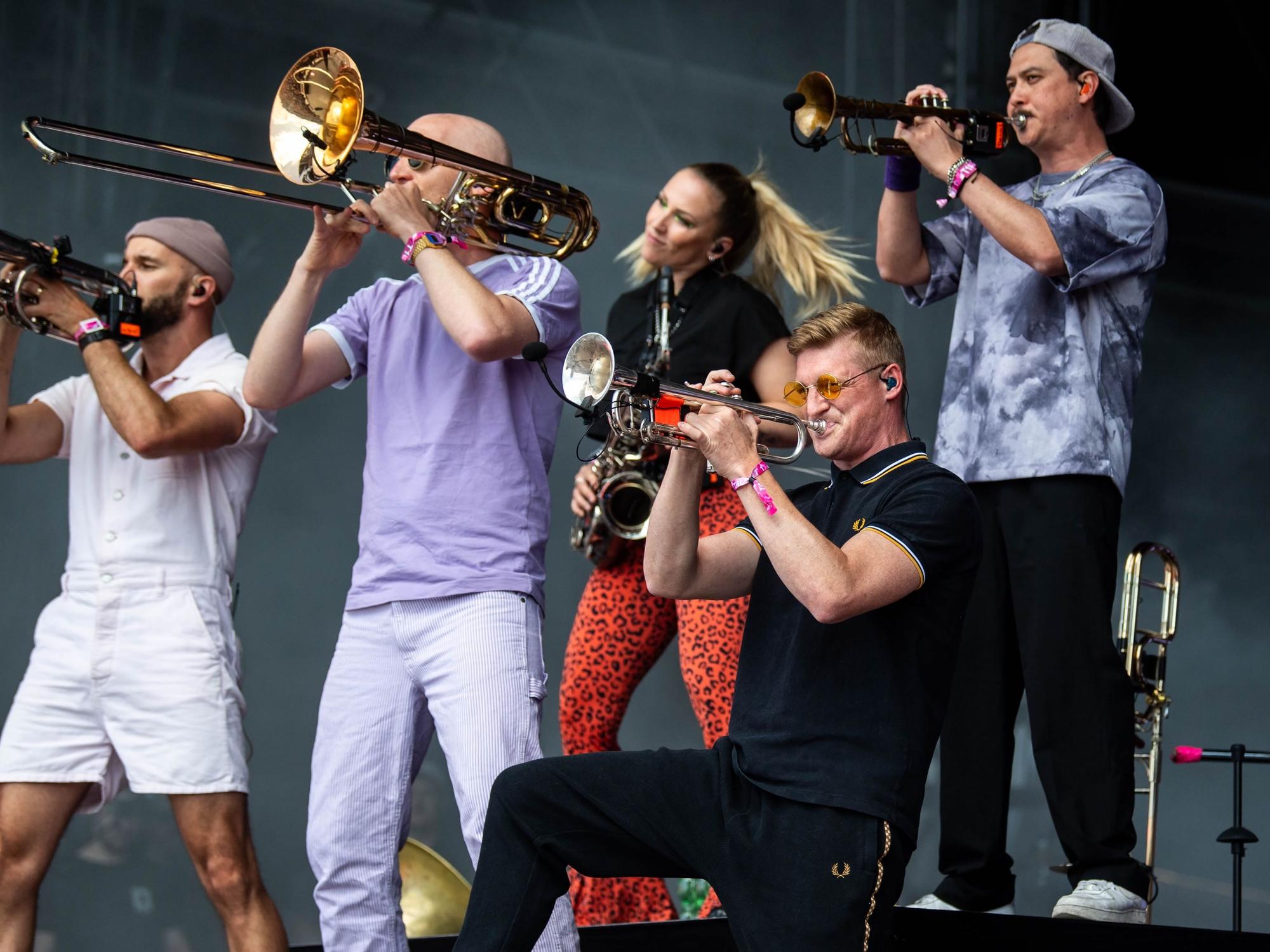 Bunt angezogenen Musikerinnen und Musiker der Band Querbeat stehen auf einer Bühne und spielen Posaune, Trompete und Saxophon.