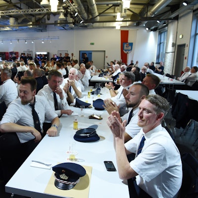 Zahlreiche Feuerwehrleute sitzen in einer Veranstaltungshalle an langen Tischreihen beisammen.