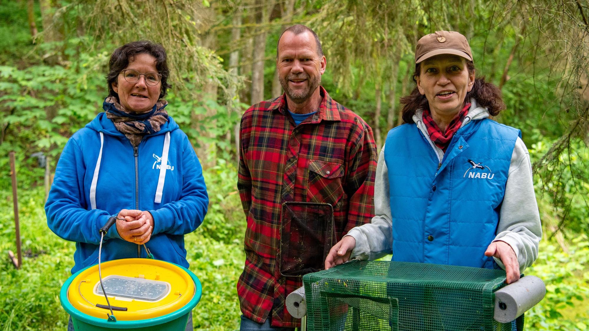 Kerstin Jonke (l.), Ulrich Pohl und Marion Zöller vom Naturschutzbund Euskirchen stehen mit Eimer, Kescher und Reuse an einem Teich. Um sie herum ist Grün.