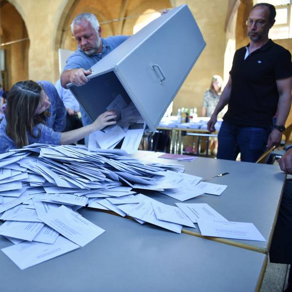 Eine Wahlurne wird von einem Mann geleert. Um den Tisch mit den Wahlzetteln stehen Wahlhelfer.