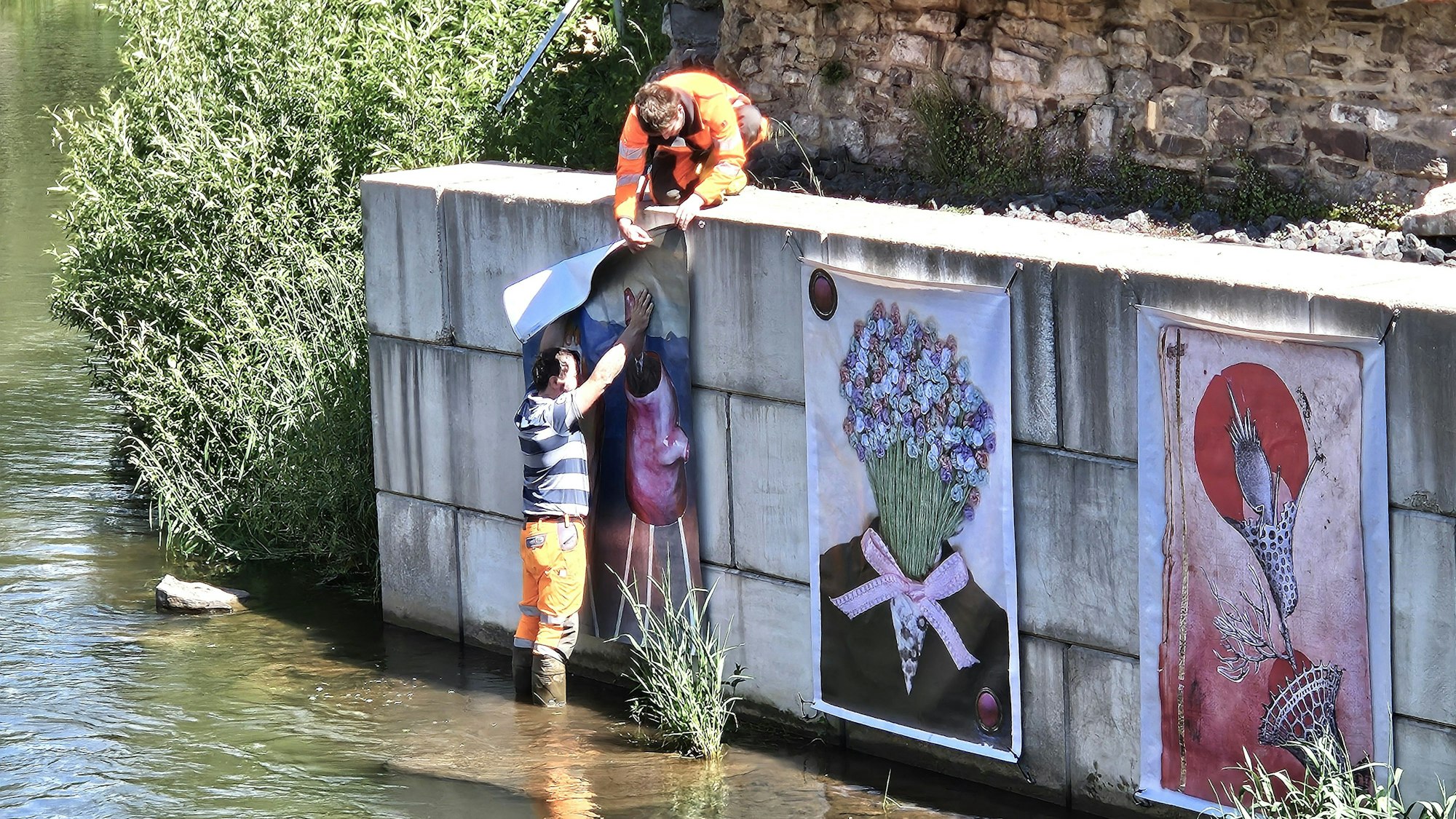 In Gemünd hängen Helfer Kunstwerke an die Uferbefestigungsmauer der Urft.