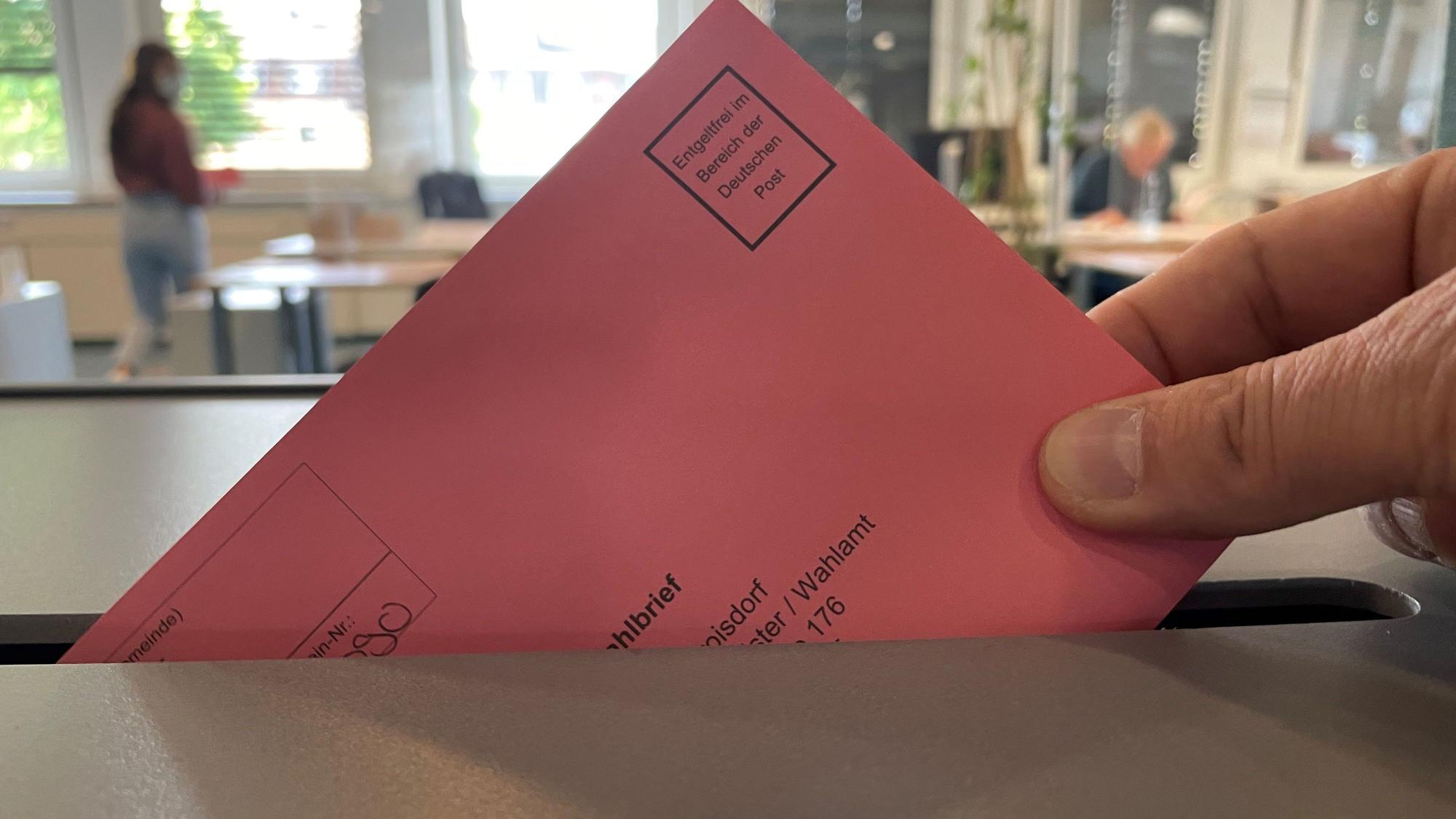 Eine Hand lässt einen roten Wahlbrief in eine Urne fallen.