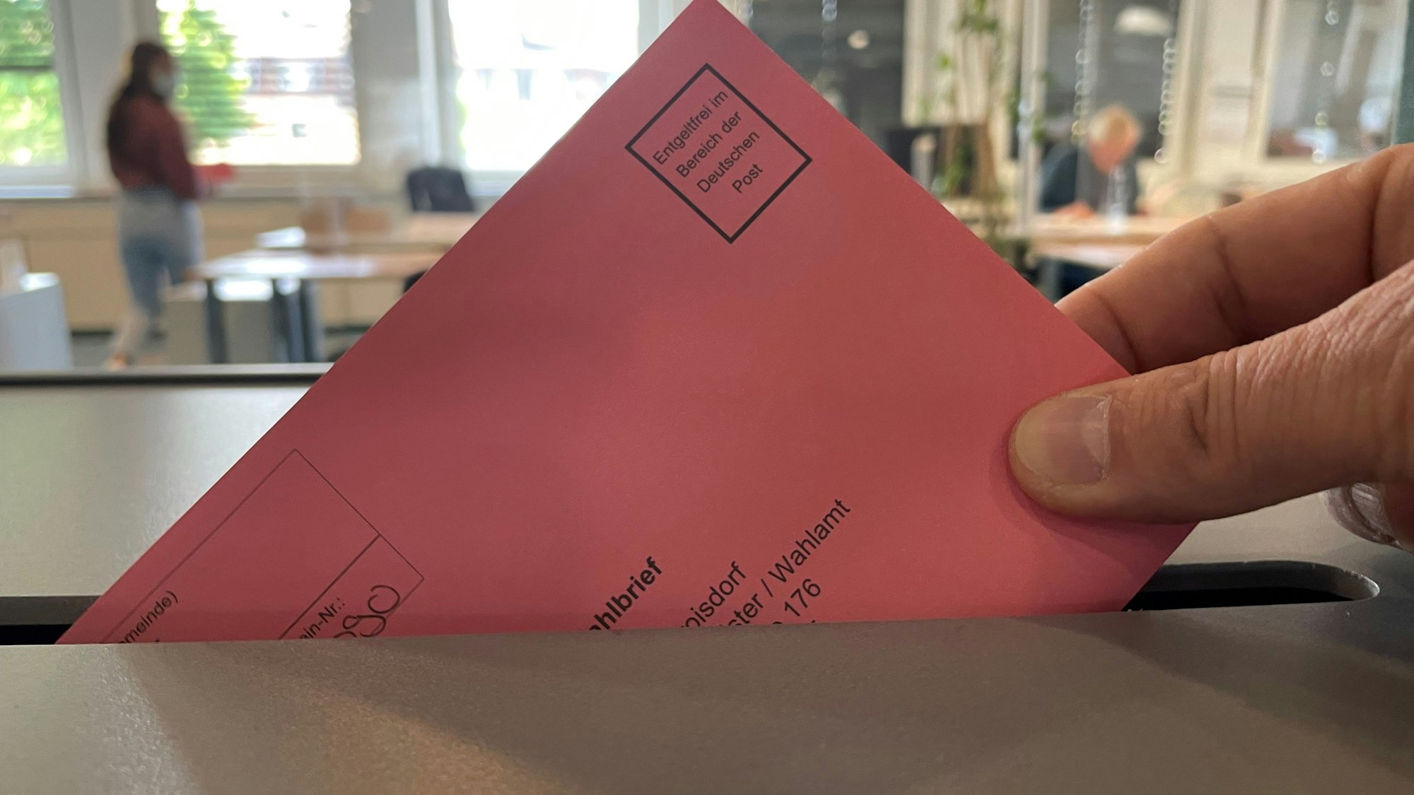 Eine Hand lässt einen roten Wahlbrief in eine Urne fallen.
