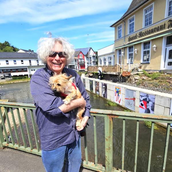 Kurator Jürgen A. Roder trägt seinen Hund auf dem Arm und steht auf einer Brücke vor den Kunstwerken.
