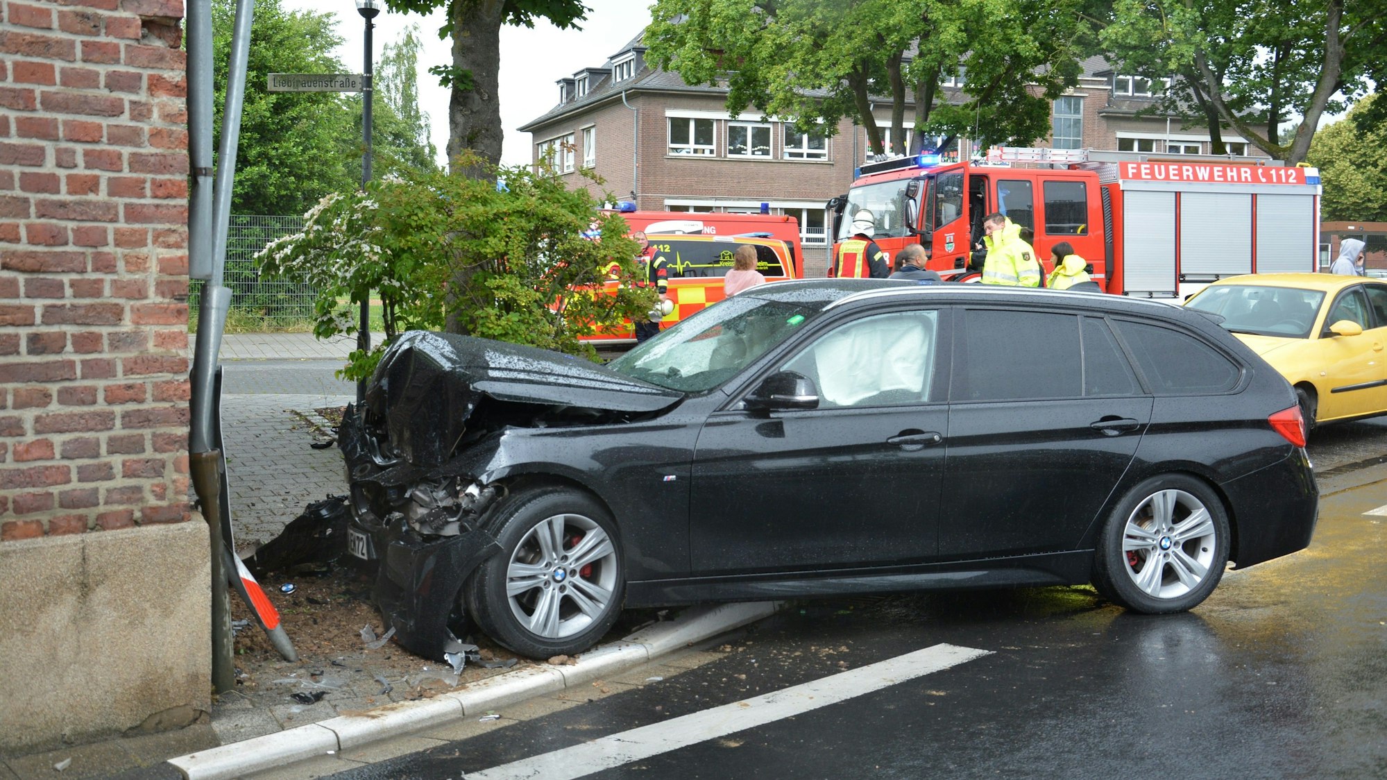 Ein Auto mit zerbeulter Motorhaube und weiteren Schäden steht vor einer Hauswand. Im Hintergrund Feuerwehr- und Rettungsfahrzeuge.