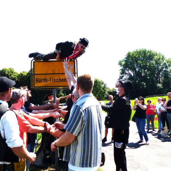 Ein junger Mann mit Hut und Schal klettert über das Ortseingangsschild von Hürth-Fischenich. Ein Empfangskomitee will ihn auffangen.