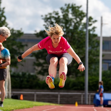 Ein Weitspringerin mitten im Sprung. Sportwarte kontrollieren die Absprungmarkierung.