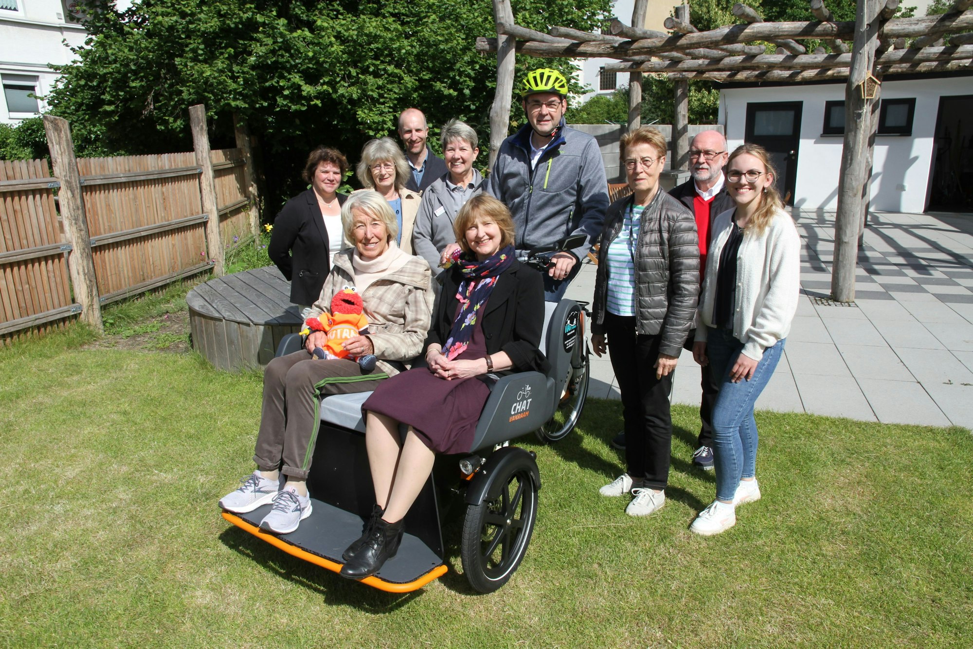 Zwei Frauen sitzen in einer Fahrradrikscha, eine Menschengruppe umringt sie. Einer von ihnen trägt einen Fahrradhelm.