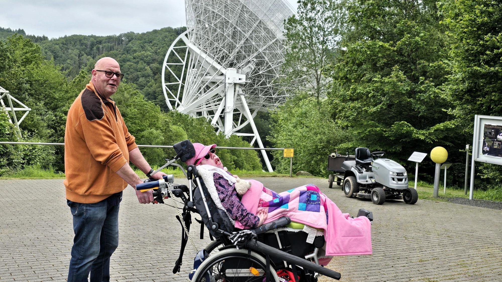 Ein Mann hält den Rollstuhl fest, in dem seine Tochter sitzt. Die beiden stehen vor dem Radioteleskop in Effelsberg.