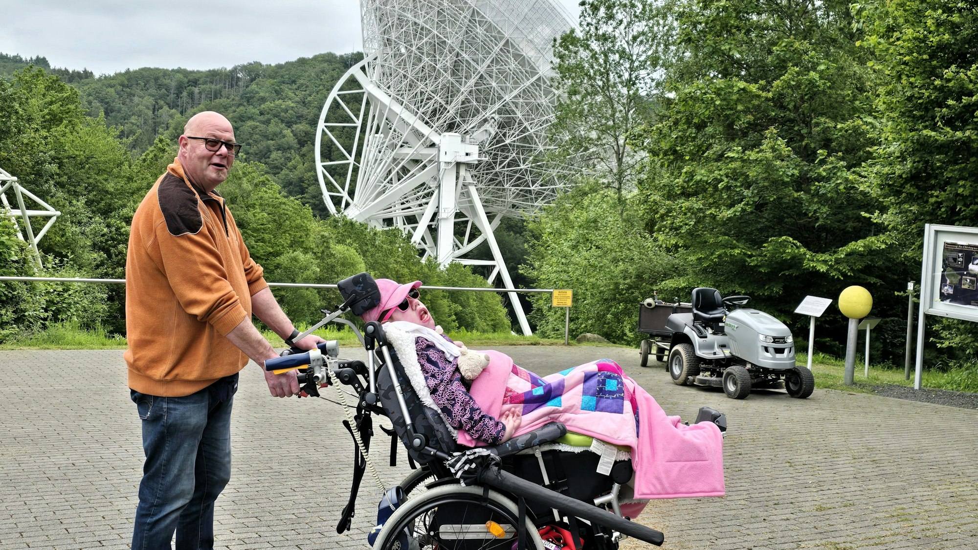 Ein Mann hält den Rollstuhl fest, in dem seine Tochter sitzt. Die beiden stehen vor dem Radioteleskop in Effelsberg.