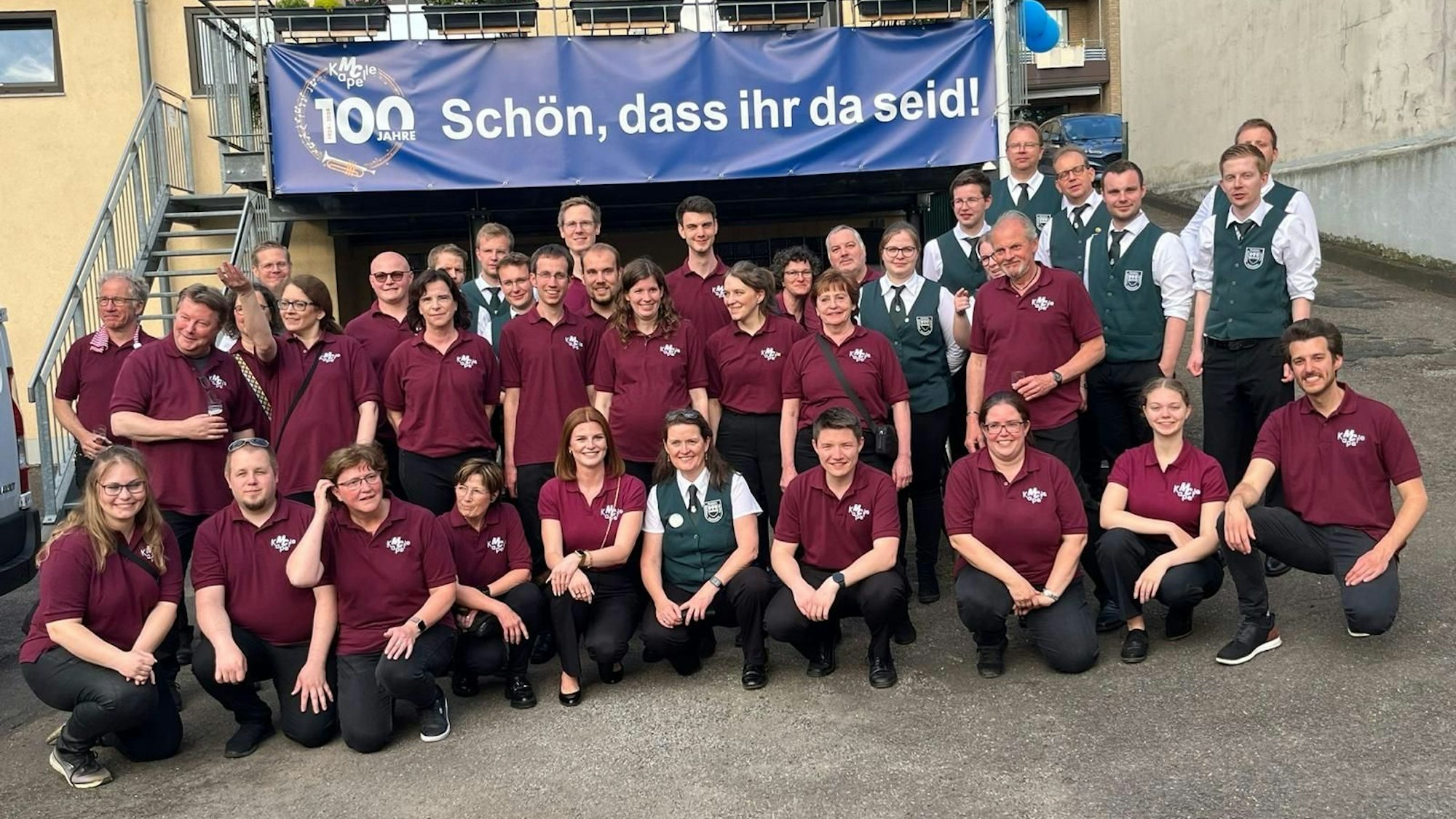 Eine Gruppe posiert in Dreier-Reihen vor einem Plakat auf dem eine 100 prangt und der Satz "Schön, dass ihr da seid!".