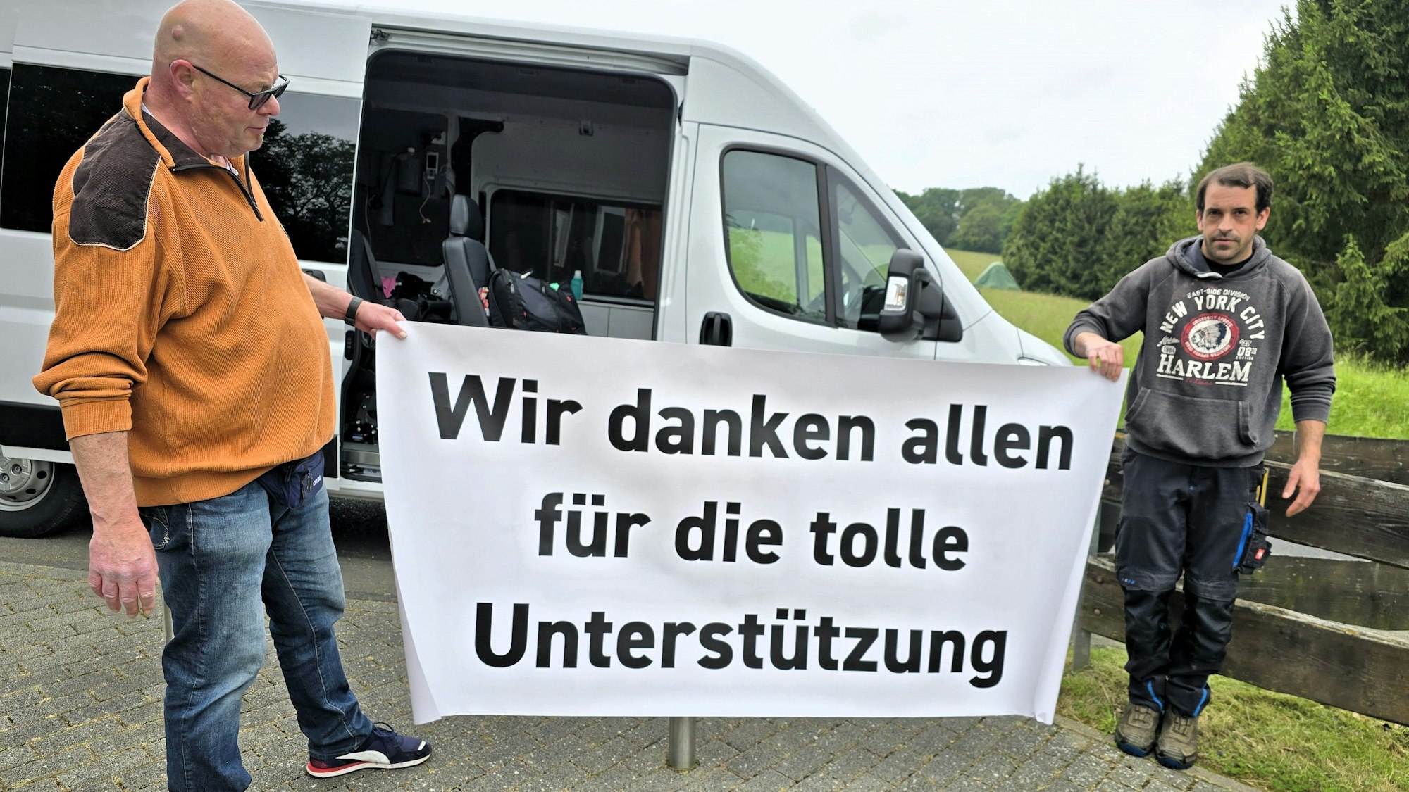 Zwei Männer stehen an einem weißen Transporter und halten ein weißes Transparent mit schwarzer Aufschrift in den Händen. Darauf steht: „Wir danken alle für die tolle Unterstützung“.