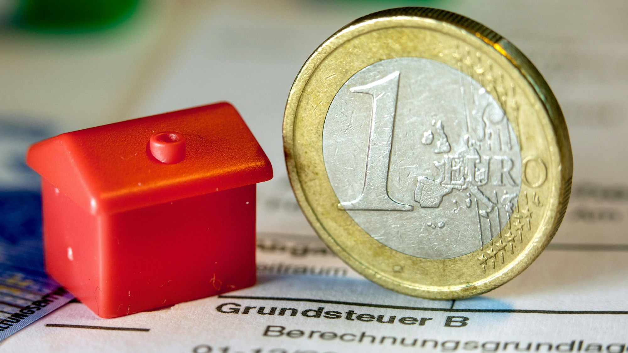 Eine Euro-Geldmünze sowie ein Spielzeughaus stehen auf einem Abgabenbescheid für die Entrichtung der Grundsteuer.