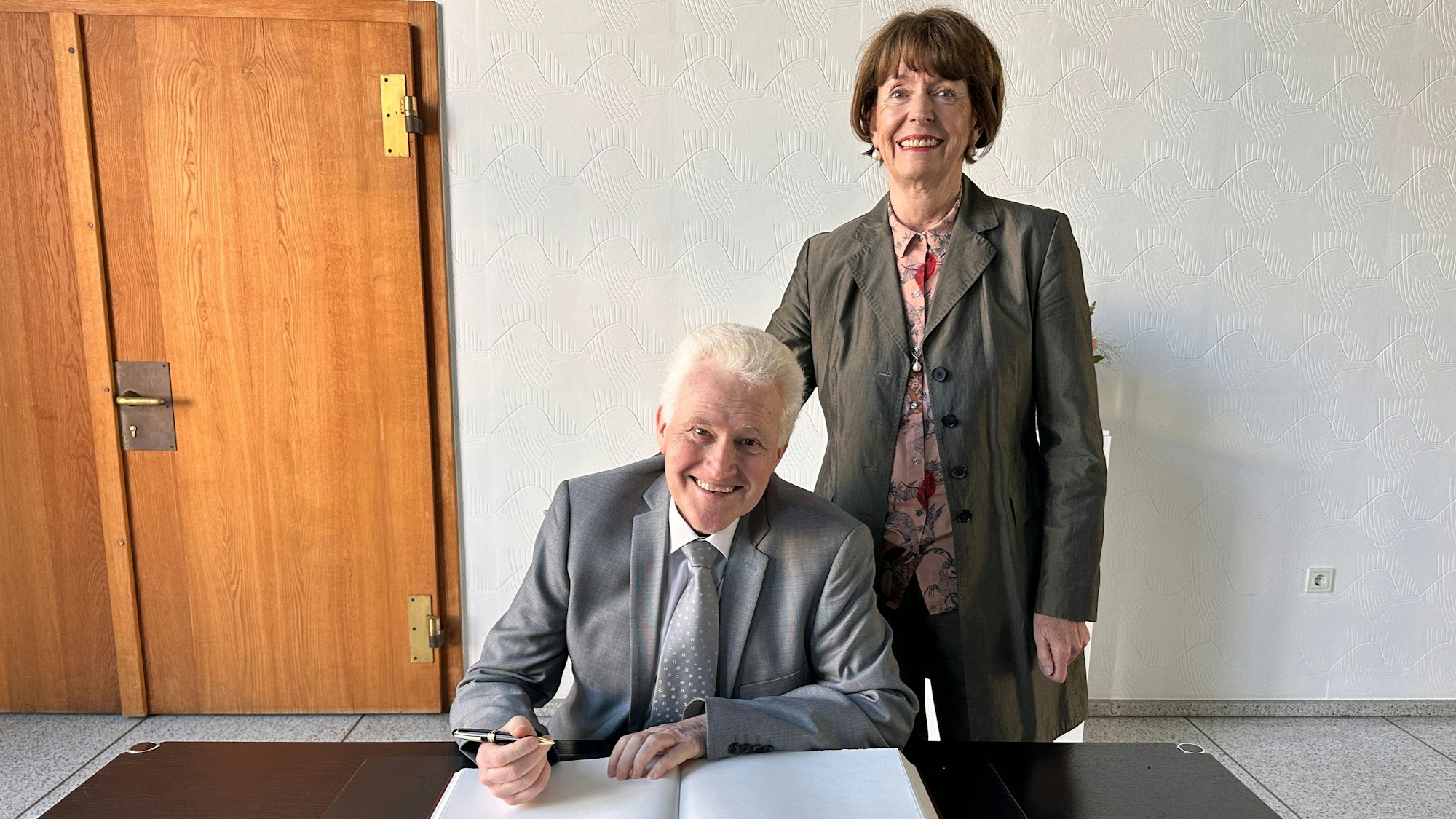 Ein grauhaariger Mann in Anzug und Krawatte sitzt an einem Tisch, Füller in der Hand, vor einem großen Buch. Neben ihm steht eine Frau im Anzug.