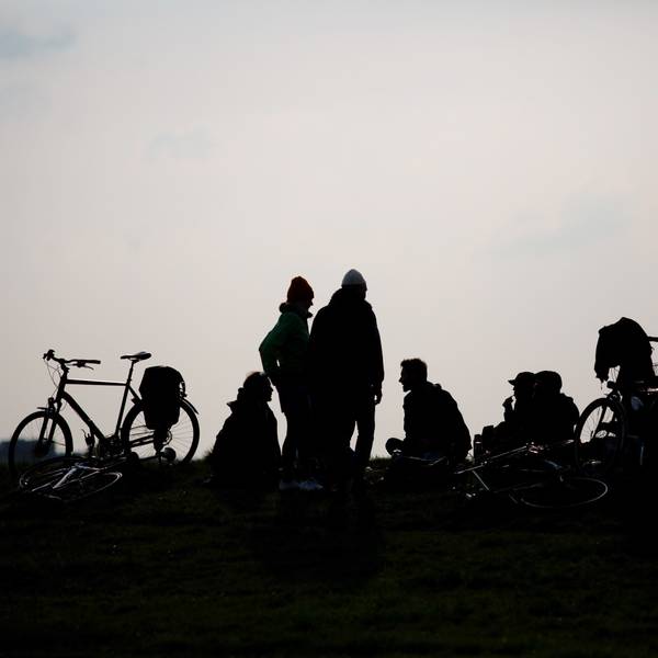 Junge Menschen stehen und sitzen auf dem Boden, Fahrräder neben sich. Das Foto ist gegen die Sonne aufgenommen, ihre Silhouetten sehen wie Schattenrisse aus.