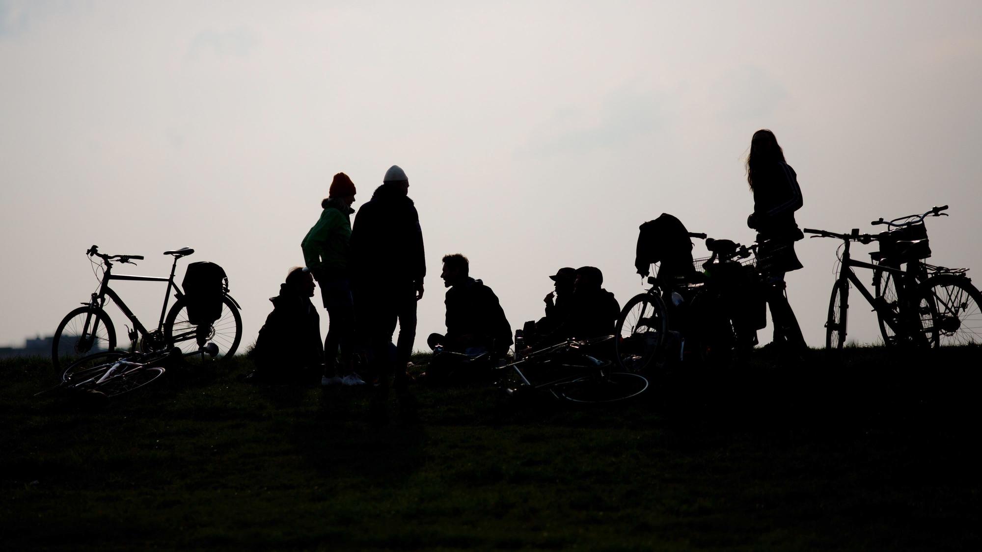 Junge Menschen stehen und sitzen auf dem Boden, Fahrräder neben sich. Das Foto ist gegen die Sonne aufgenommen, ihre Silhouetten sehen wie Schattenrisse aus.