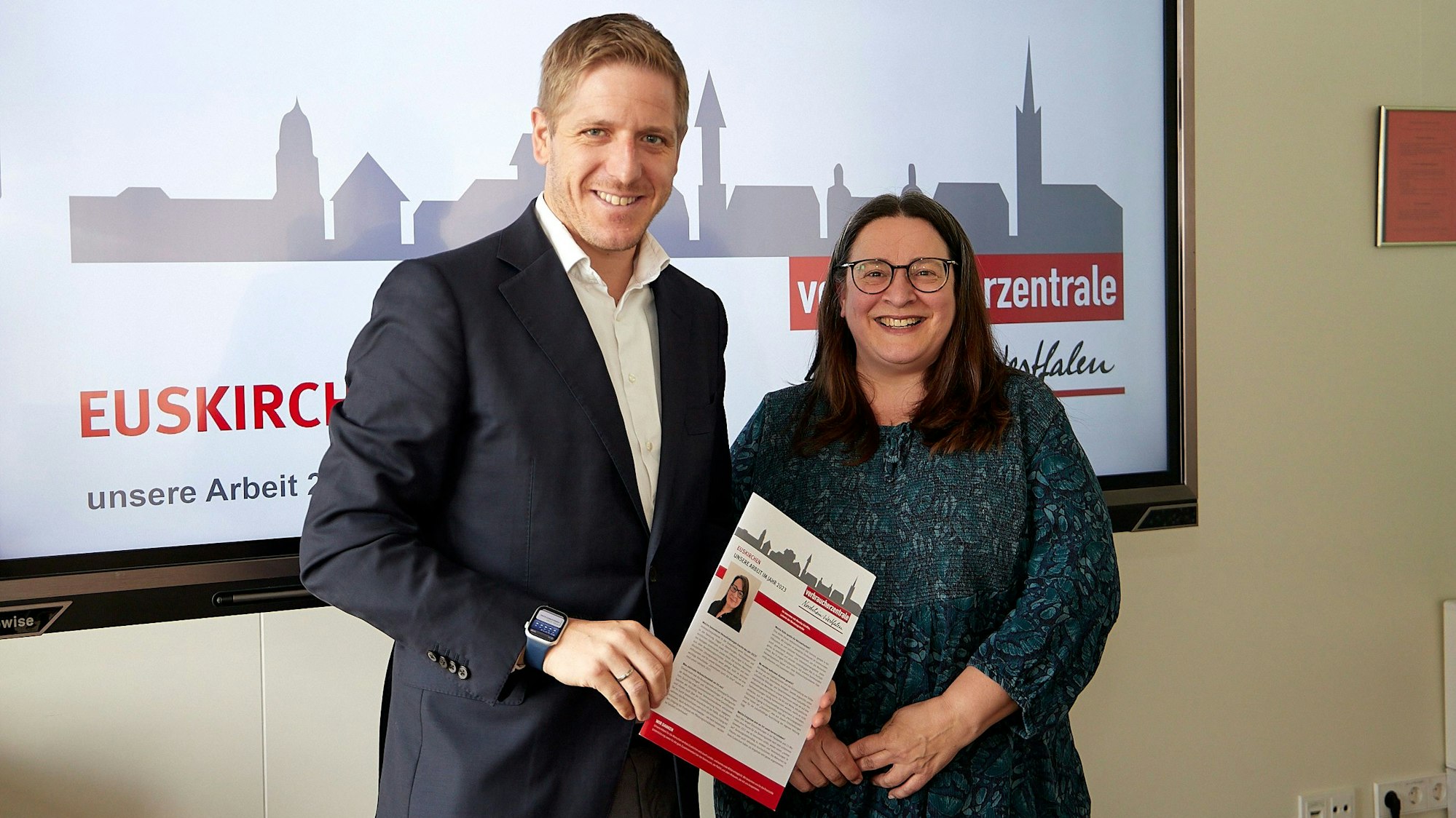 Landrat und Markus Ramers und Monika Schiffer, die Leiterin der Beratungsstelle, stehen vor einem Bildschirm mit dem Logo der Verbraucherzentrale Euskirchen. Ramers hält den Jahresbericht in der Hand.