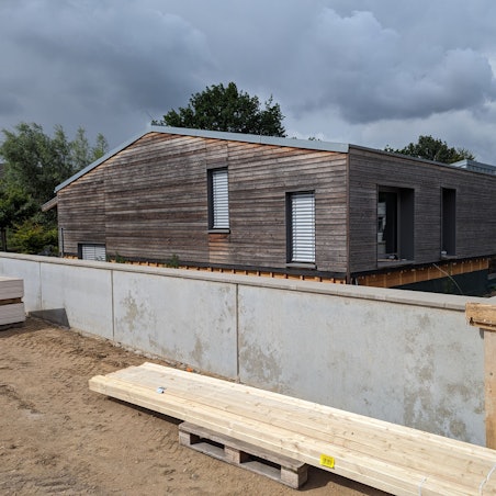 Auf dem Foto ist eine Kita in Holzbauweise zu sehen. Um sie herum wird eine Mauer gegen Überflutungen gebaut.
