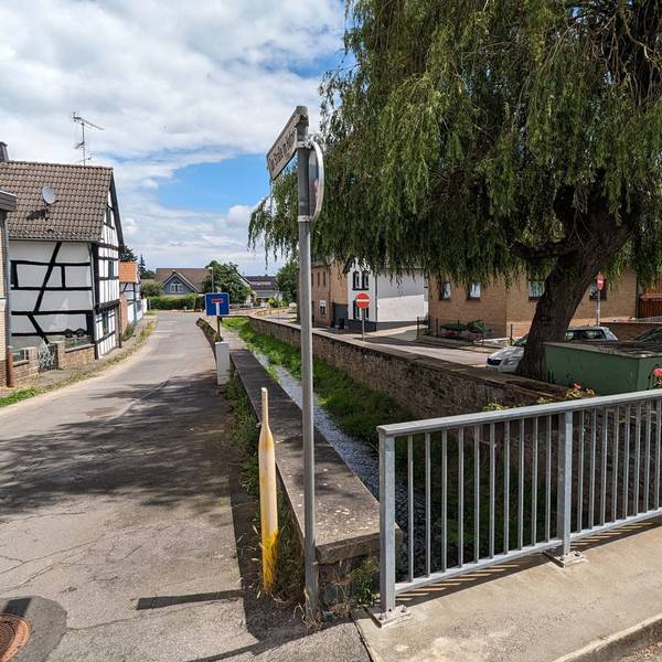 Im Bereich der Rehgasse in Kommern verläuft der Bleibach zwischen alten Ufermauern. Vorne ist das Geländer einer Brücke zu sehen.