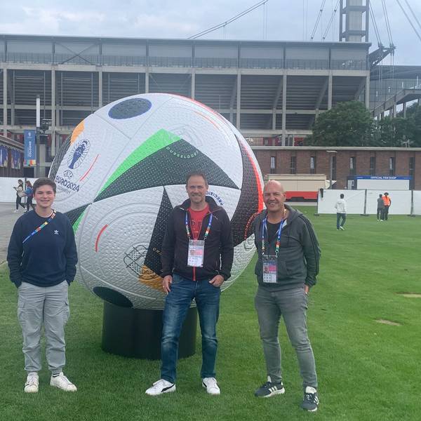 Das Bild zeigt die drei freiwilligen Helfer vor dem Rhein-Energie-Stadion in Köln. Sie stehen vor einem überdimensionalen Spielball.
