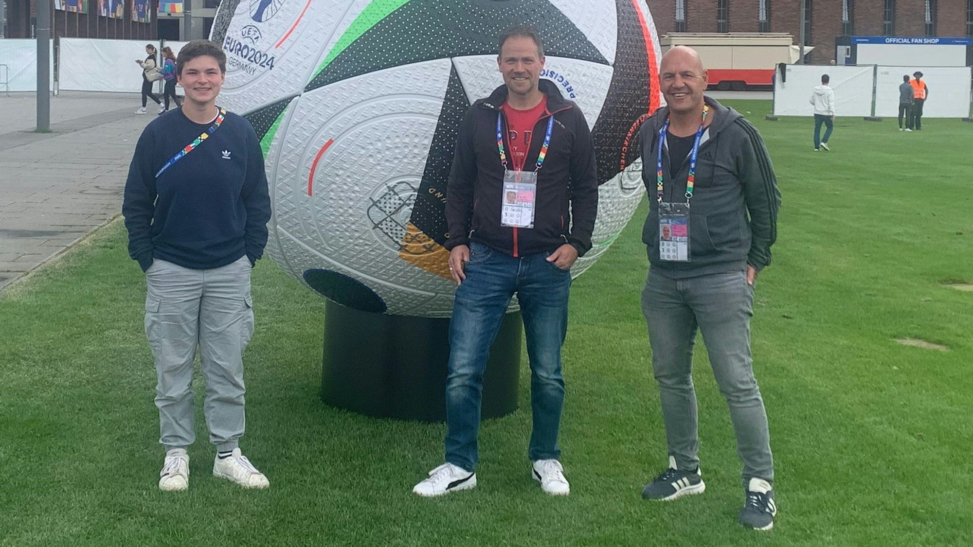 Das Bild zeigt die drei freiwilligen Helfer vor dem Rhein-Energie-Stadion in Köln. Sie stehen vor einem überdimensionalen Spielball.