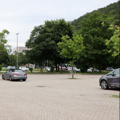 Auf dem Parkplatz am Sittardweg stehen ein paar Autos. Im Hintergrund, zwischen den Baumkronen, kommt der fast fertig gestellte Neubau an der Trierer Straße heraus.