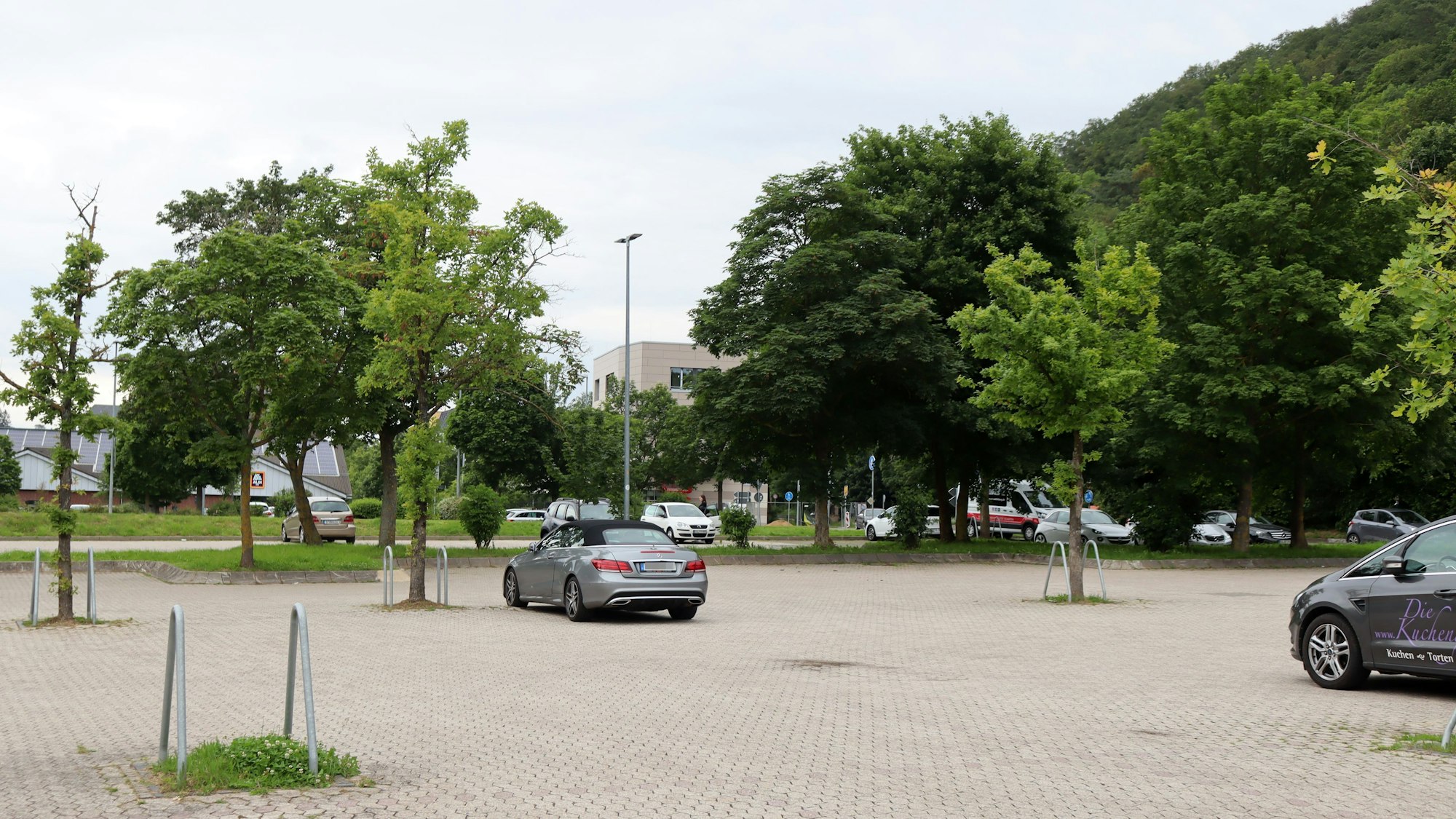 Auf dem Parkplatz am Sittardweg stehen ein paar Autos. Im Hintergrund, zwischen den Baumkronen, kommt der fast fertig gestellte Neubau an der Trierer Straße heraus.