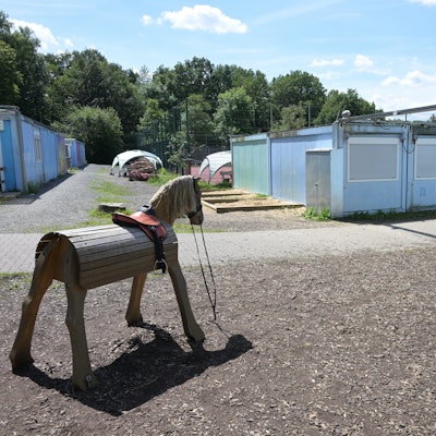 Rechts und links stehen Containerbauten, im Vordergrund zwei hölzerne Spielpferde.