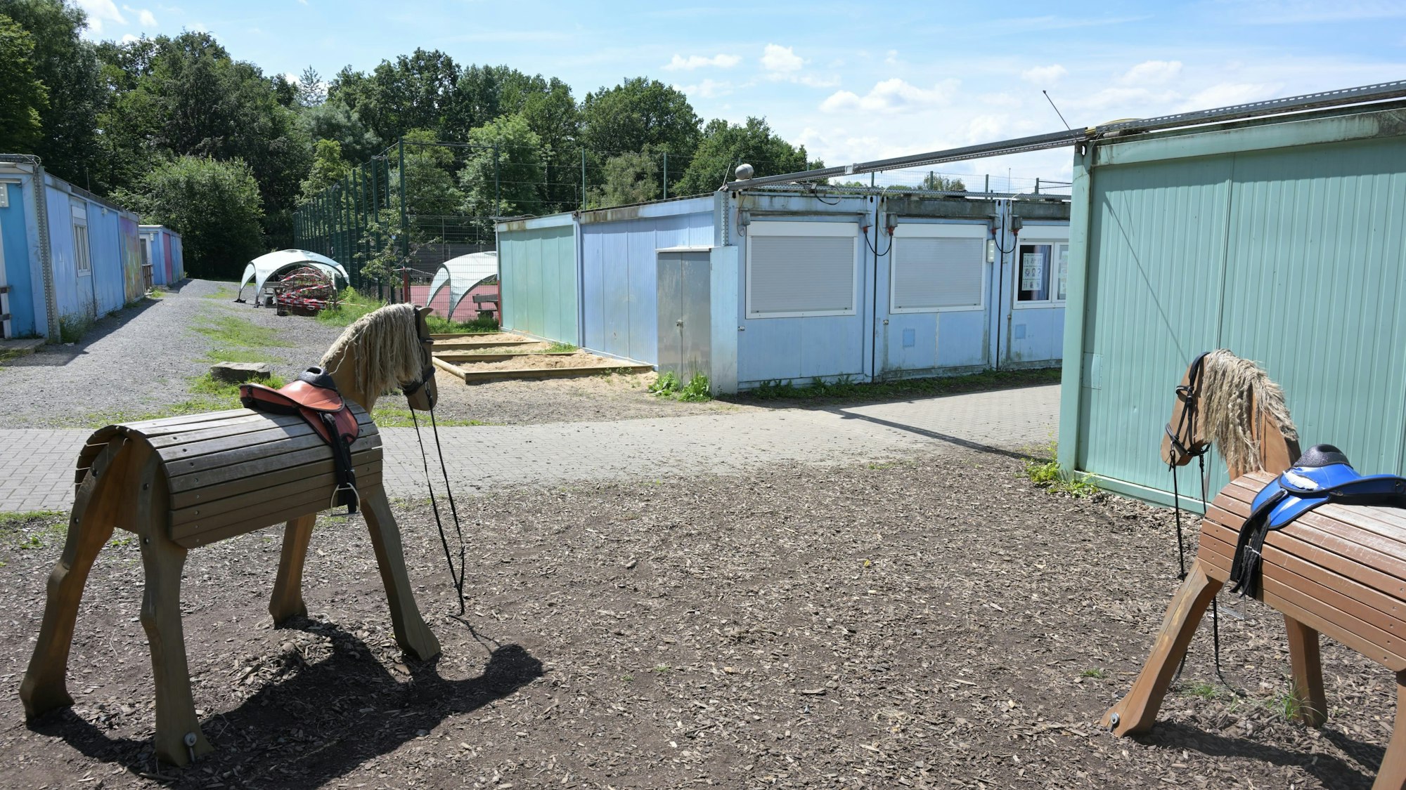 Rechts und links stehen Containerbauten, im Vordergrund zwei hölzerne Spielpferde.