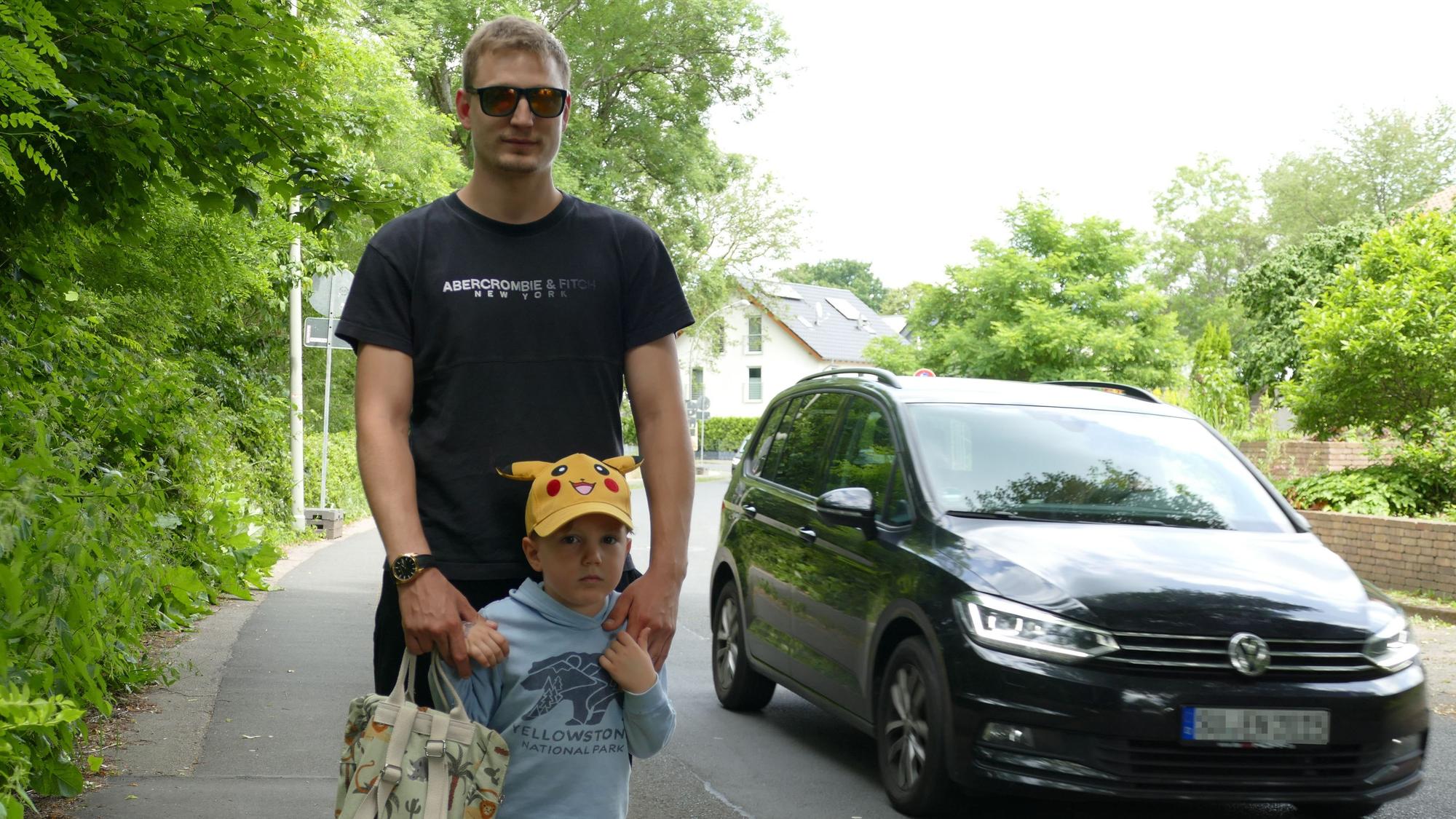 Ein Mann und ein kleiner Junge auf dem Bürgersteig, daneben fährt ein Auto vorbei.