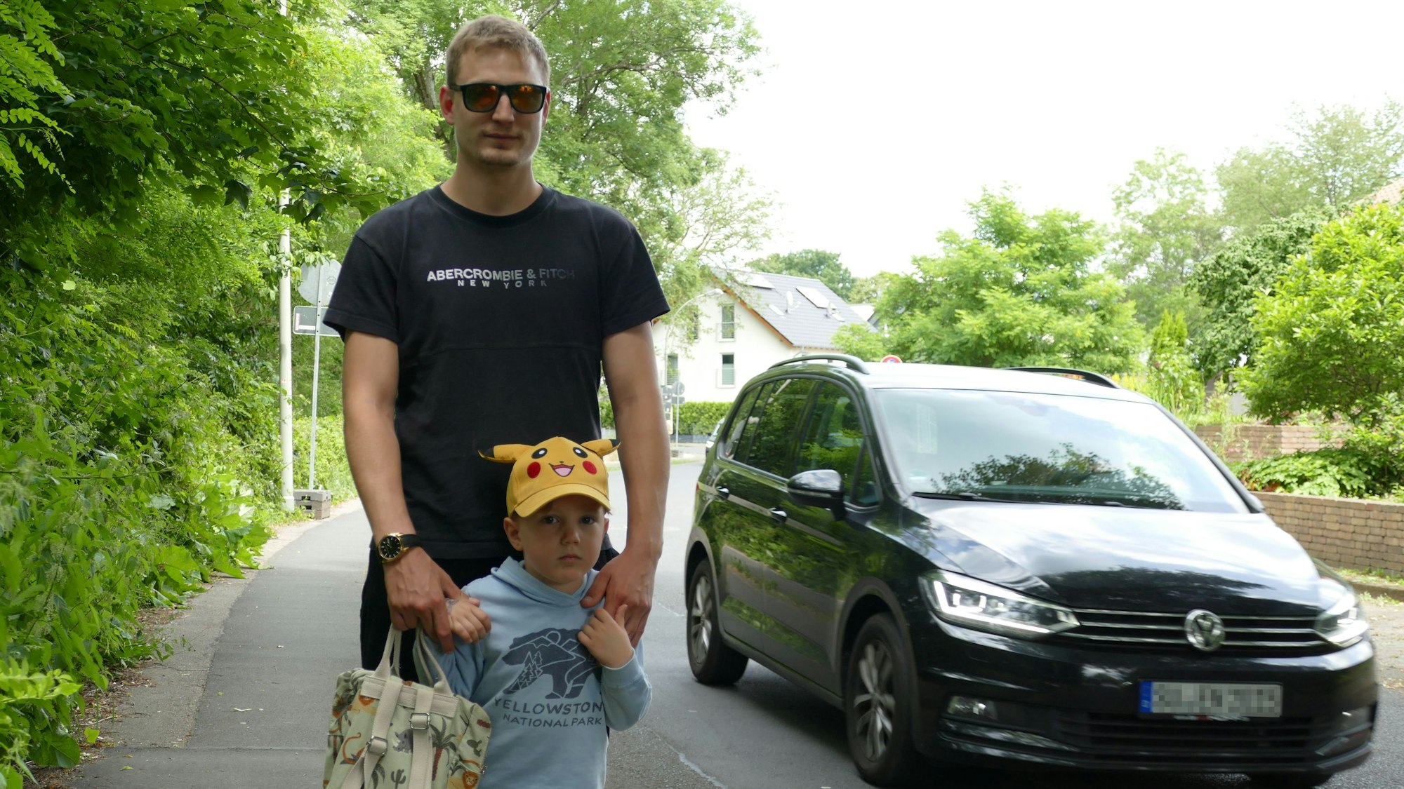 Ein Mann und ein kleiner Junge auf dem Bürgersteig, daneben fährt ein Auto vorbei.