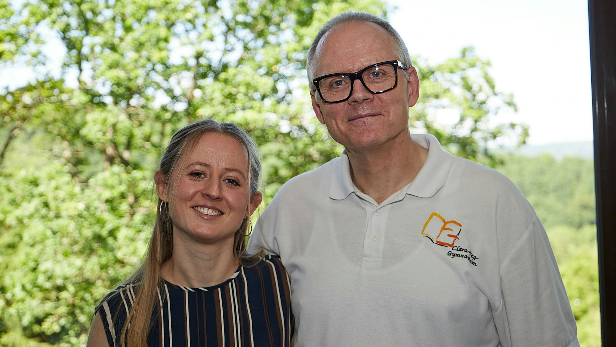 Rudolf Berens und seine Tochter Hannah stehen zusammen vor einem Fenster, im Hintergrund sind grüne Bäume zu sehen.