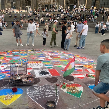 Passanten am Kölner Dom betrachten ein Kreidebild, das die Fahnen vieler Länder zeigt.