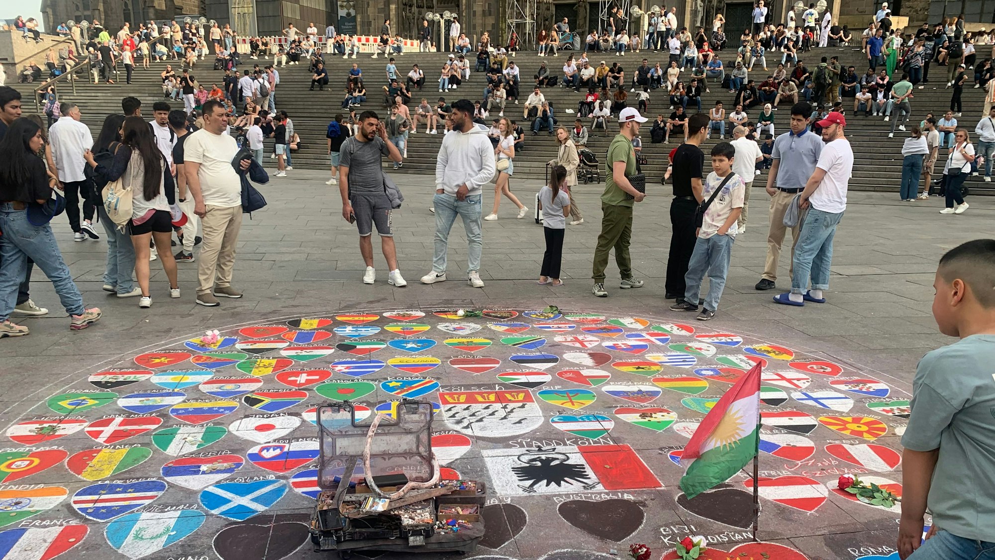 Passanten am Kölner Dom betrachten ein Kreidebild, das die Fahnen vieler Länder zeigt.
