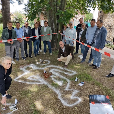Elke Pistor und Ralf Kramp befinden sich in einem mit Flatterband abgesperrten Bereich, in dem die Umrisse einer imaginären Leiche sowie dargestellte Blutspuren zu sehen sind. Organisatoren und Sponsoren des Krimifestivals schauen ihnen zu.