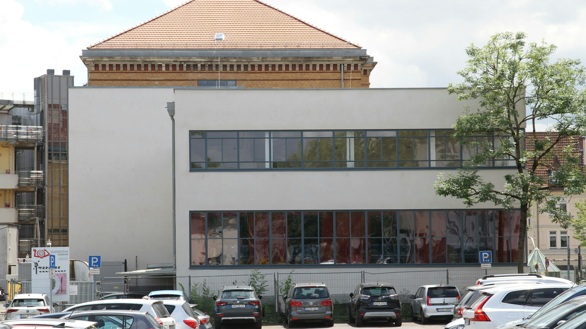 Ein graues Haus mit Flachdach und großen Fensterflächen an einem Parkplatz