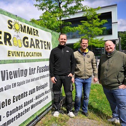 Timo Bay, Frank Hewel und Mike Bierögel stehen vor einem großen Banner, das auch den Sommer Biergarten mit Public-Viewing aller EM-Spiele hinweist.