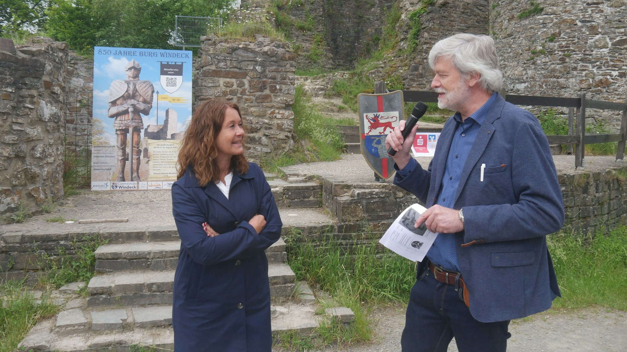 Ein Mann mit Mikrofon spricht mit einer Frau, sie stehen vor einer Burgruine.