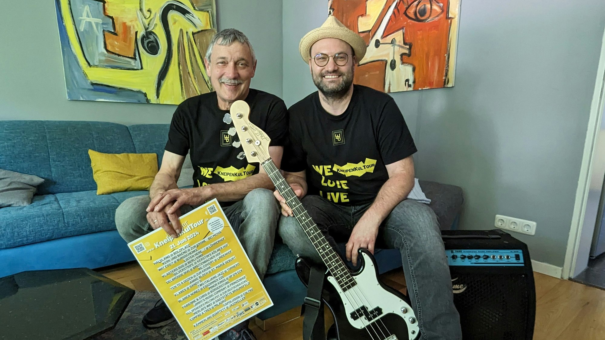 Zwei Männer mit Plakat, Bassgitarre und einem Verstärker auf einem Sofa.