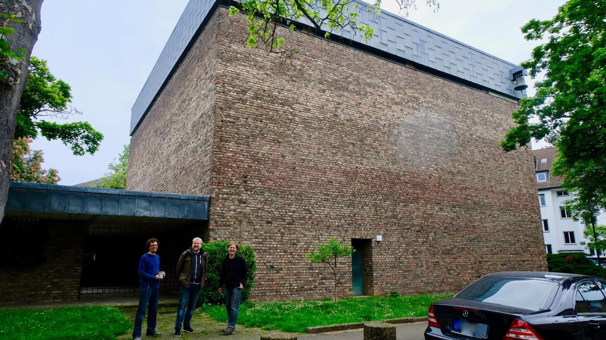 Vor einem hohen würfelförmigen Beton-Gebäude stehen drei Männer.