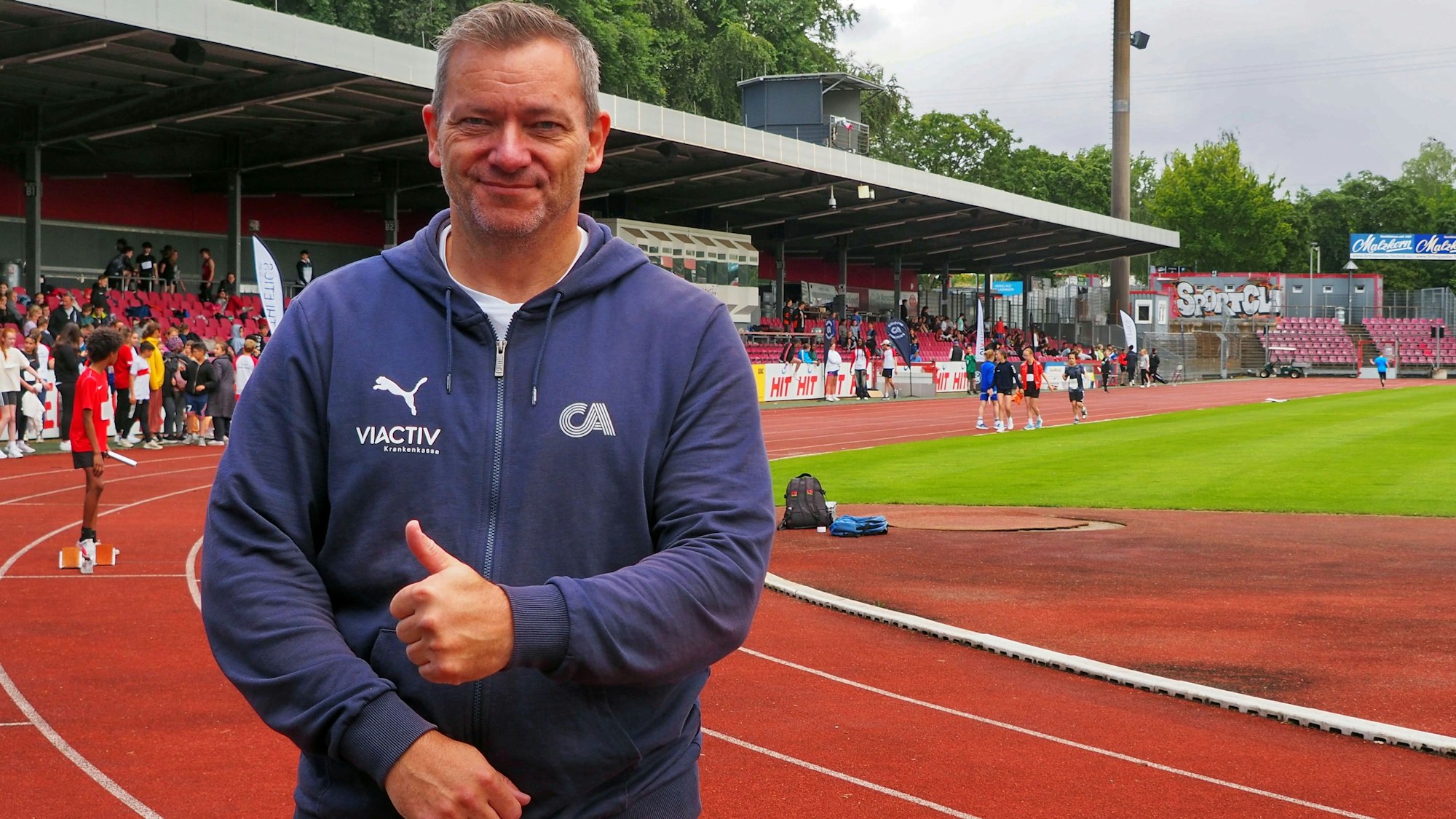 Ein Mann in Jeans und Sportjacke zeigt die Daumen-Hoch-Geste. Er steht auf der Laufbahn in einem Stadion.