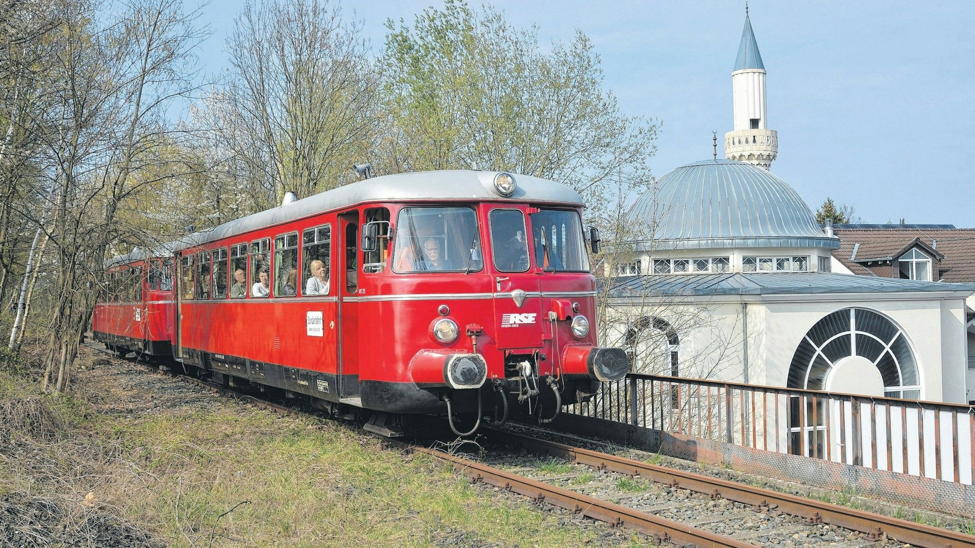 Ein roter, altmodischer Schienenbus befährt ein Gleis. Rechts sieht man das Minarett einer Moschee.