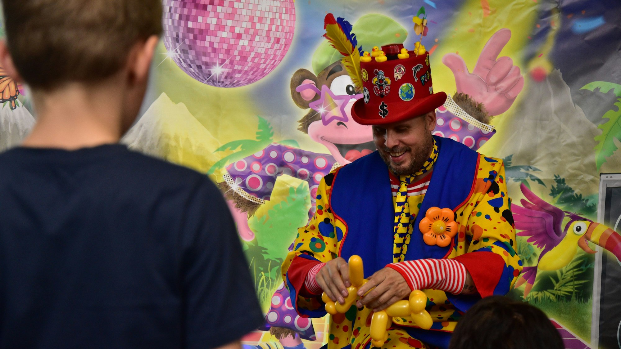 Ein bunt kostümierter Mann formt für die jungen Besucherinnen und Besucher des neuen Indoorspielplatzes Monkey Town Figuren aus Luftballons.